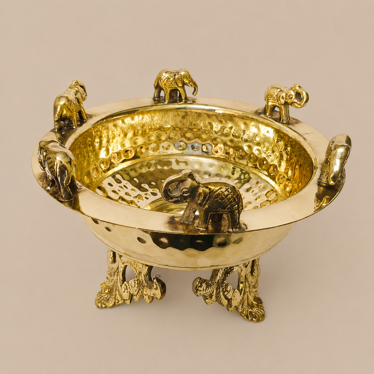 A decorative brass Urli bowl with elephant motifs, featuring a hammered finish on the interior, a hammered smooth exterior, three elephant feet as legs, and small elephant figures sitting on the rim, photographed against a solid beige background.