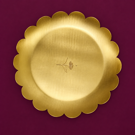 A Brass round gold scalloped-edge serving platter with a brushed metallic finish and a small engraved botanical motif in the center, set against a solid deep burgundy background.