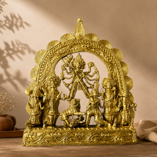 A golden Dokra Durga idol with metallic finish, displayed on a wooden surface against a warm beige wall with soft shadows.
