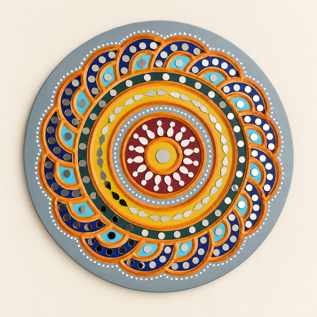 A round decorative Lippan art wall hanging featuring a colorful circular mandala pattern with concentric rings, scalloped petal-like shapes, and dotted details in blue, orange, yellow, red, green, white, and gray on a light gray base, shown against a plain beige background.
