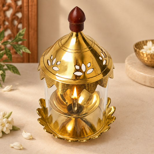 A brass Akhand jyot oil lamp with a clear glass body, decorative cutout lid, and a small flame inside, styled on a beige surface with flowers and a bowl in the background.