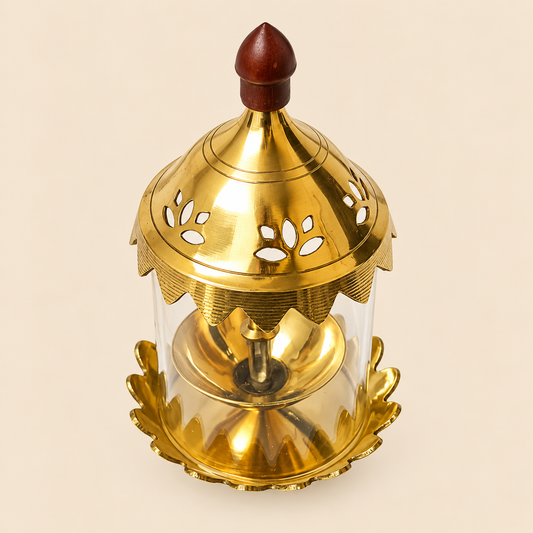 A brass Akhand jyot oil lamp with a clear glass cylinder body, featuring a polished gold-toned metal top and base with decorative cutouts and a dark reddish-brown wooden finial on top, set against a solid light beige background.
