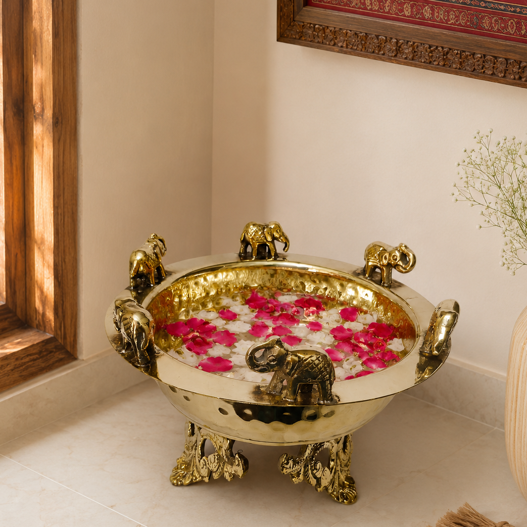 A hammered golden brass Urli with elephant-shaped handles and legs, filled with water and pink flower petals, placed on a light tiled floor in a warm indoor setting.