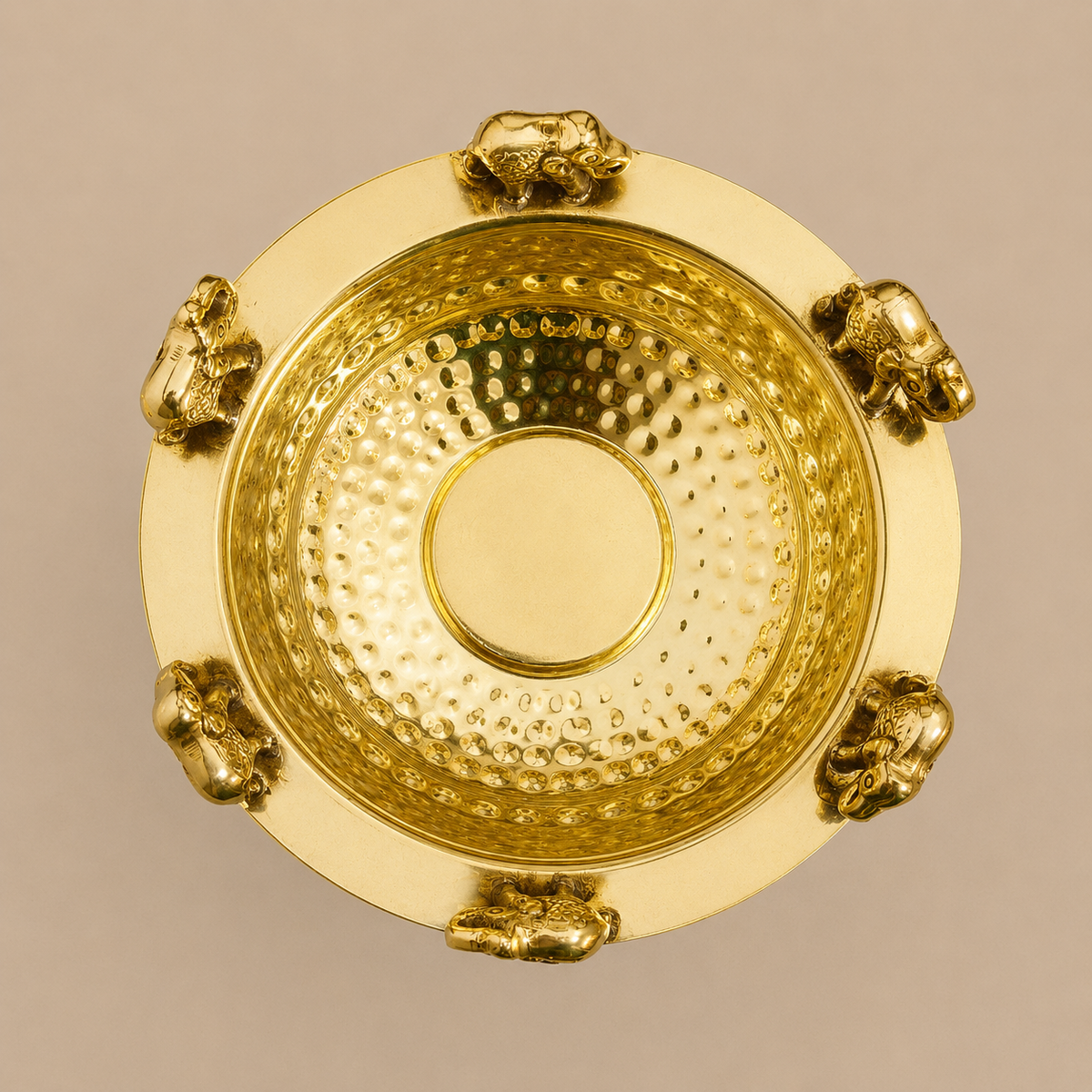 A round gold-toned brass Urli bowl with a hammered interior and a smooth outer rim, decorated with small elephant figures around the edge, shown against a plain warm beige background.