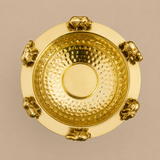 A round gold-toned brass Urli bowl with a hammered interior and a smooth outer rim, decorated with small elephant figures around the edge, shown against a plain warm beige background.