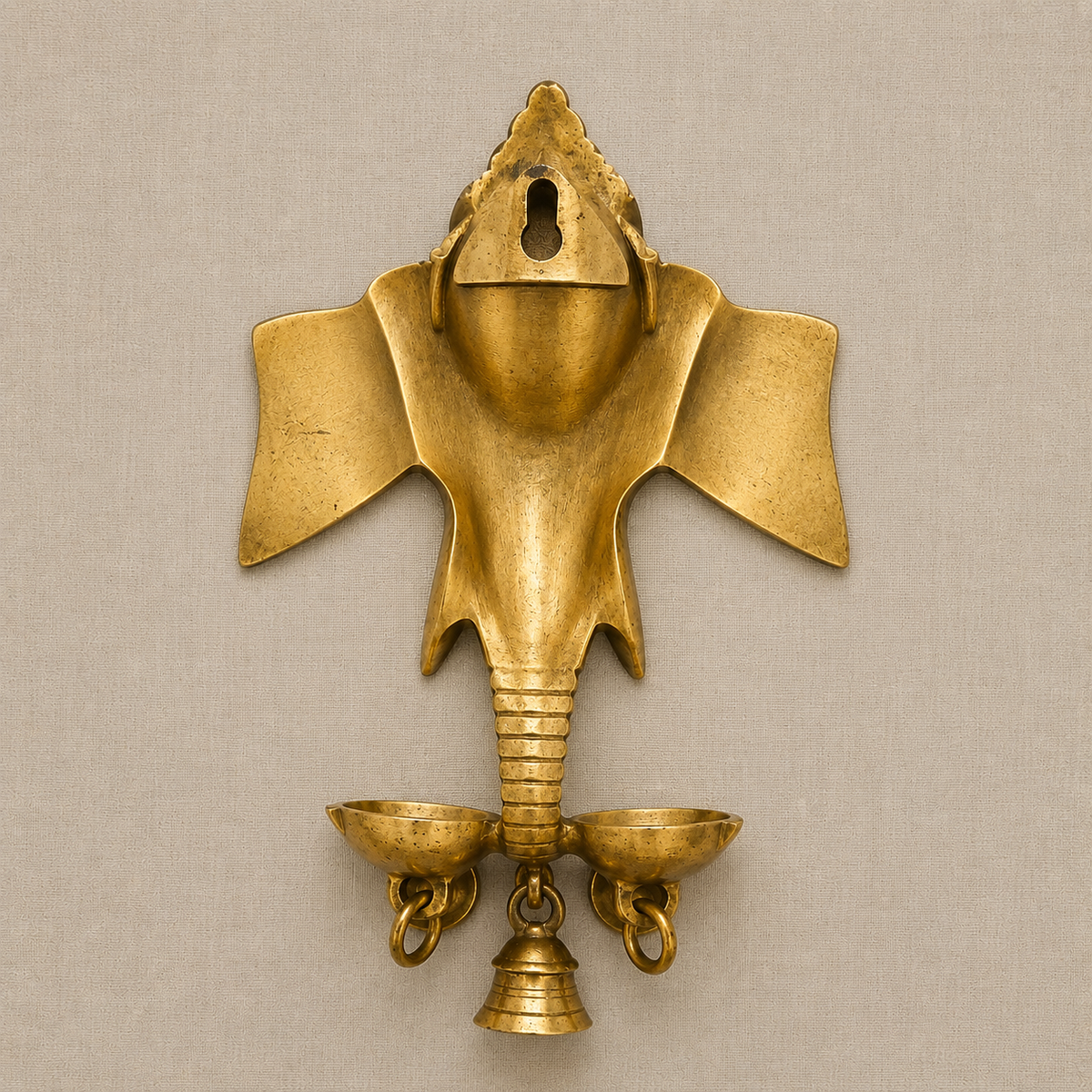 An ornate brass Ganesha hanging oil lamp with colorful stone work details and three hanging bells, set against a solid beige background.