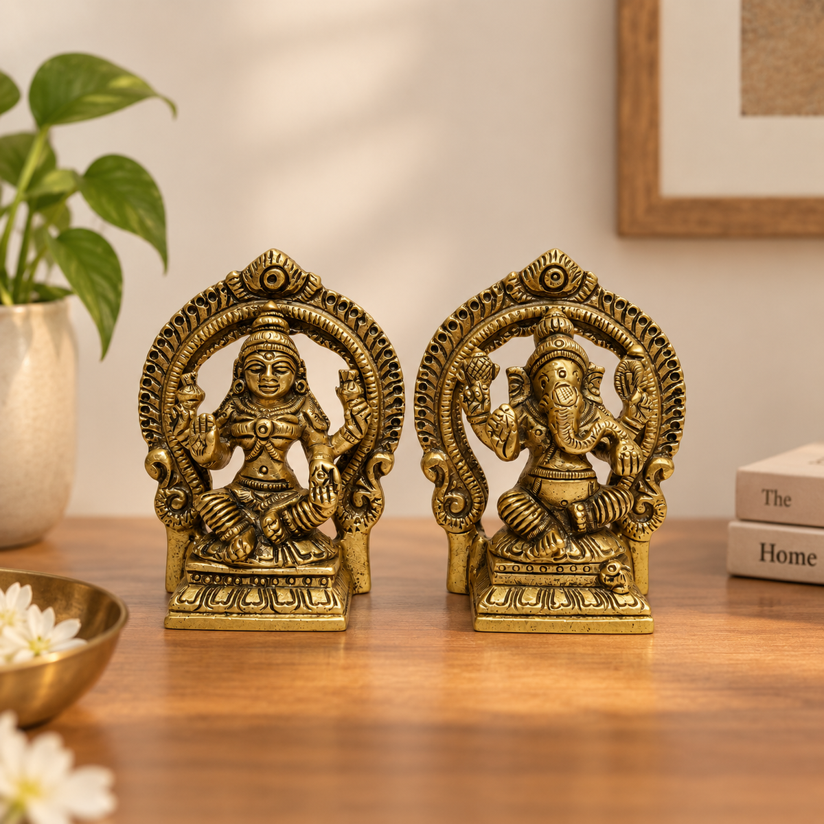 Two ornate brass Lord Ganesha and Lakshmi figurines displayed side-by-side on a wooden table, with a potted plant and books in the background.