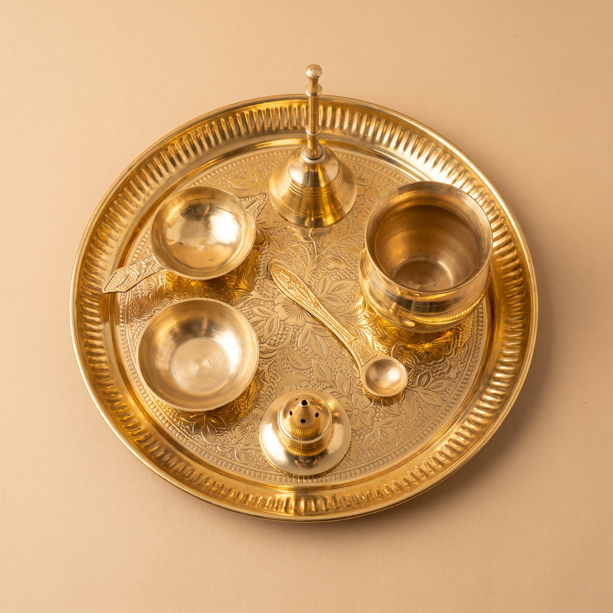 A decorative brass puja Thali set of 7, featuring a small brass kalash, bowl, diya, ritual spoon, incense holder and a bell on a Brass plate, photographed on a warm beige background.