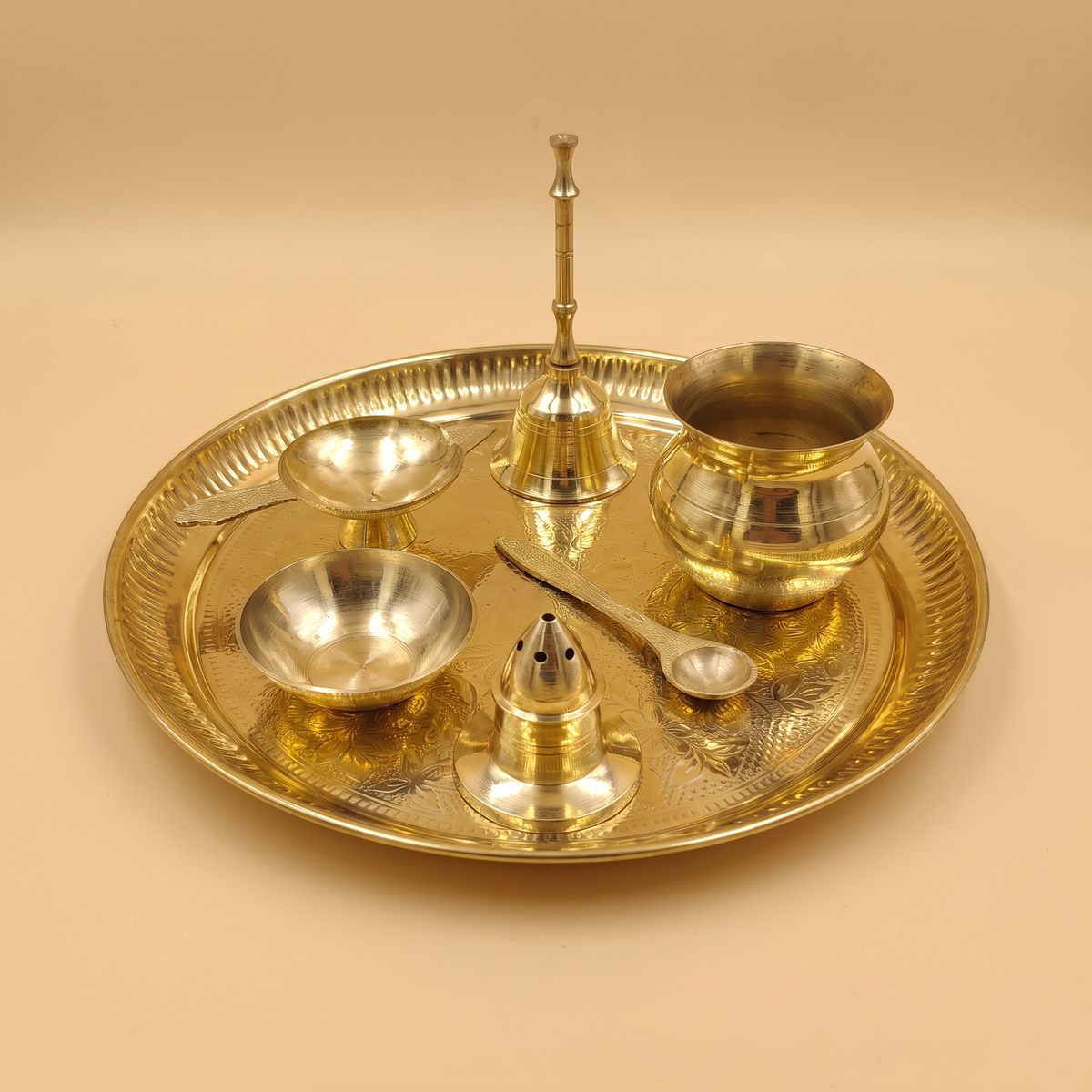 A decorative brass puja Thali set of 7, featuring a small brass kalash, bowl, diya, ritual spoon, incense holder and a bell on a Brass plate, photographed on a warm beige background.