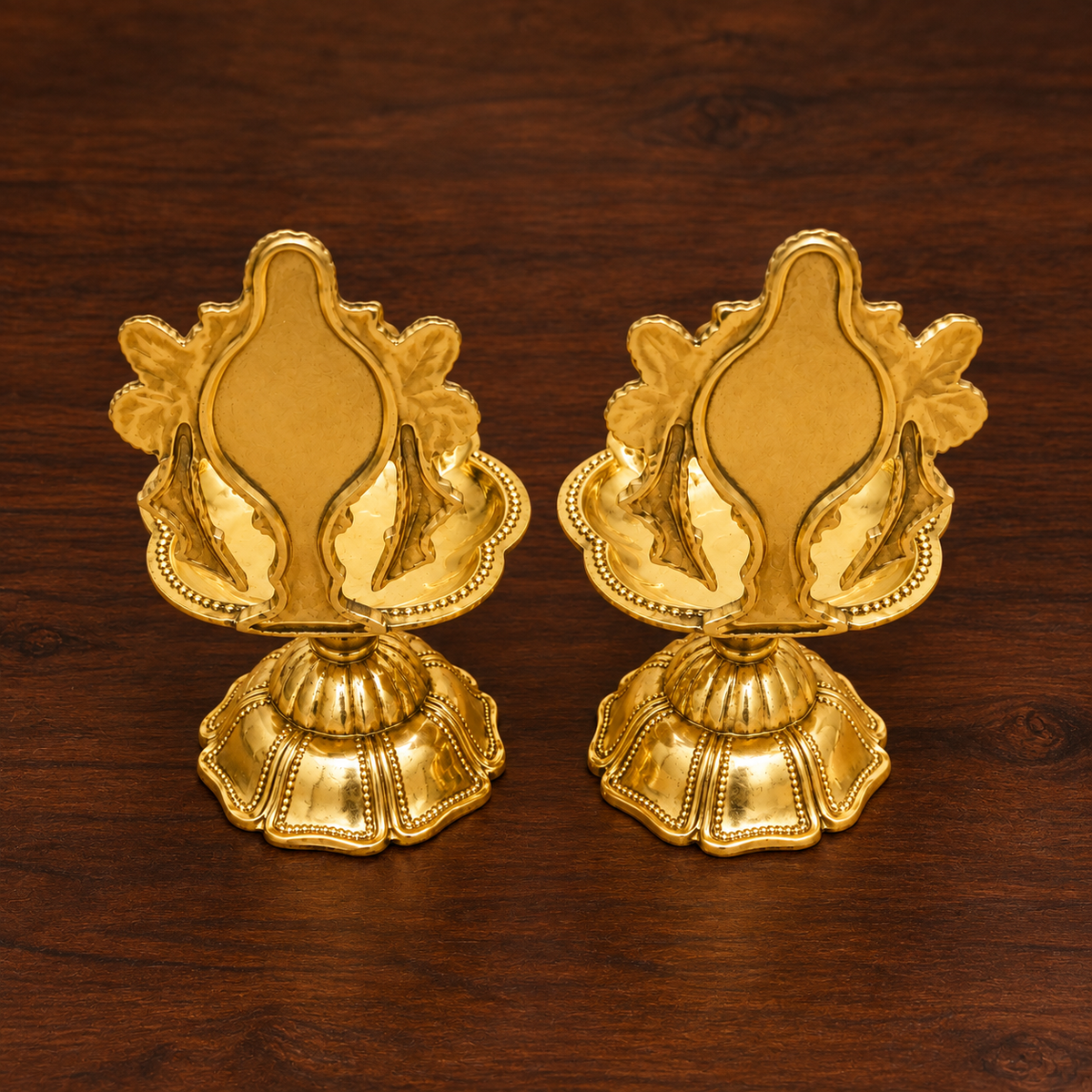 A pair of ornate brass oil lamps with decorative Shankh and Chakra finials and visible flames, placed on a wooden surface in a warm indoor setting.