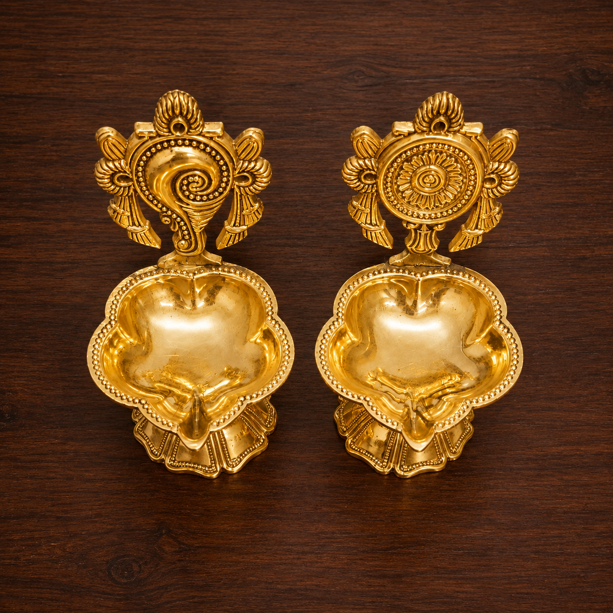 A pair of ornate brass oil lamps with decorative Shankh and Chakra finials and visible flames, placed on a wooden surface in a warm indoor setting.