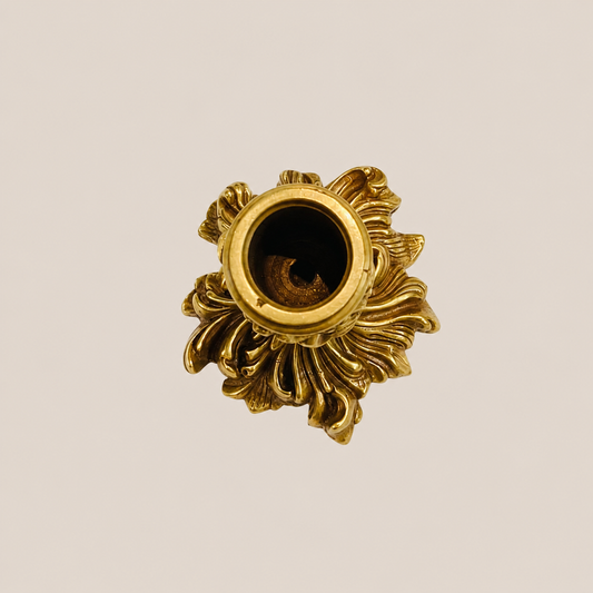An ornate gold brass candle holder with a decorative floral design, shown from a top-down view on a solid light beige background.