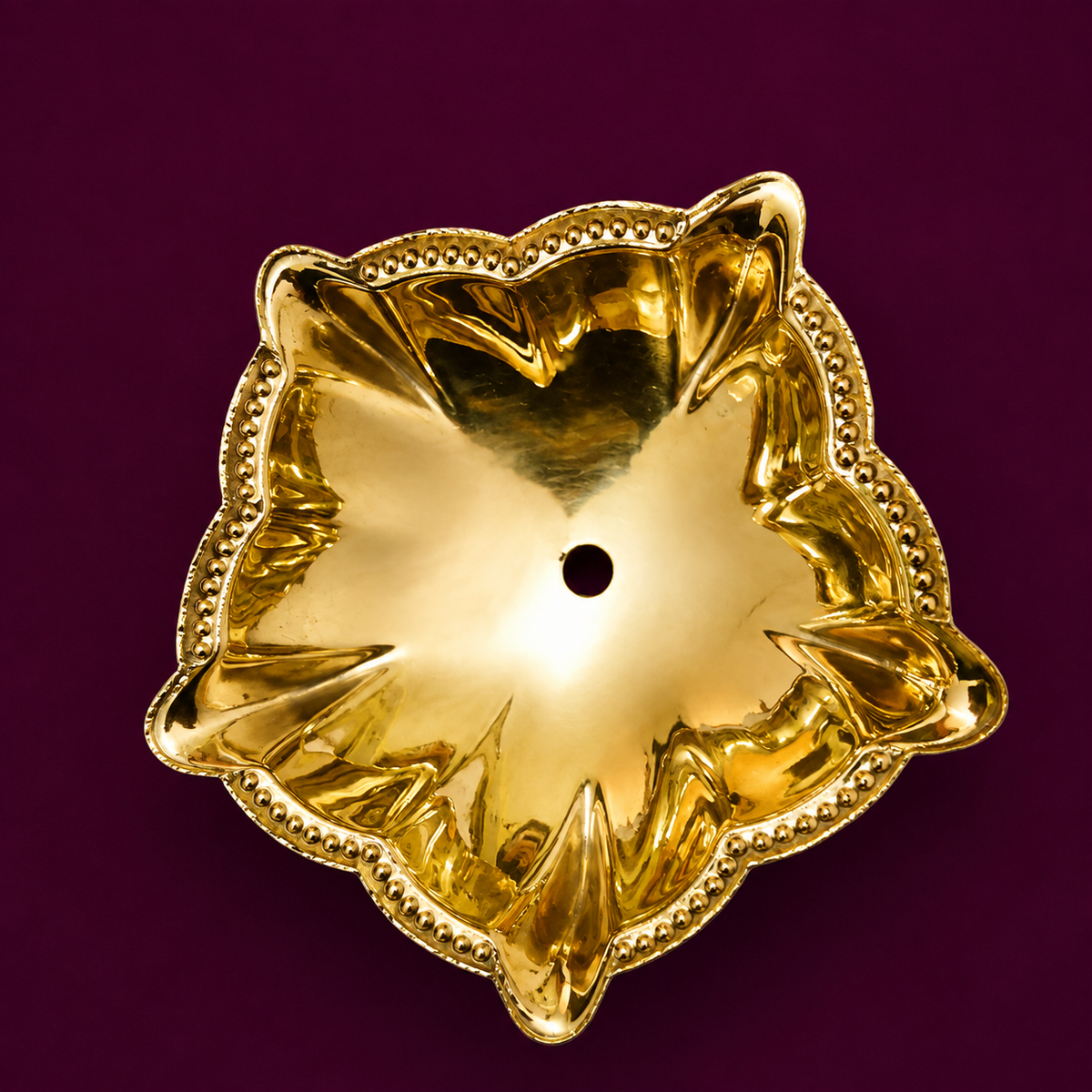 A shiny golden brass flower-shaped oil lamp, photographed from above against a solid deep purple background.
