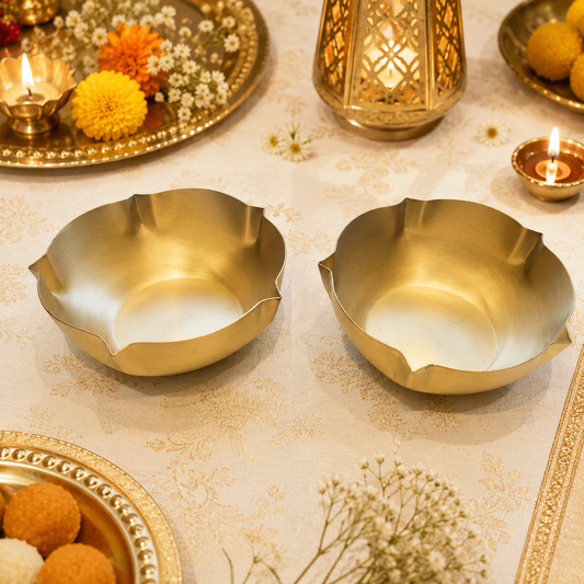 Two gold-toned brass floral shaped bowls arranged on a beige fabric table runner, surrounded by lit oil lamps, decorative flowers, and a red patterned textile.