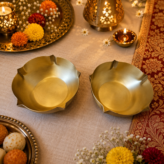 Two gold-toned brass floral shaped bowls arranged on a beige fabric table runner, surrounded by lit oil lamps, decorative flowers, and a red patterned textile.