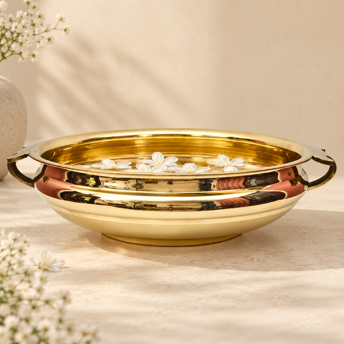 A polished golden plain brass Urli bowl with side handles, shown on a warm beige surface with small white flowers around it. The bowl is reflective and has a warm golden hue under soft lighting.