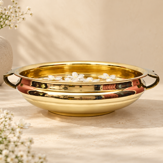 A polished golden plain brass Urli bowl with side handles, shown on a warm beige surface with small white flowers around it. The bowl is reflective and has a warm golden hue under soft lighting.