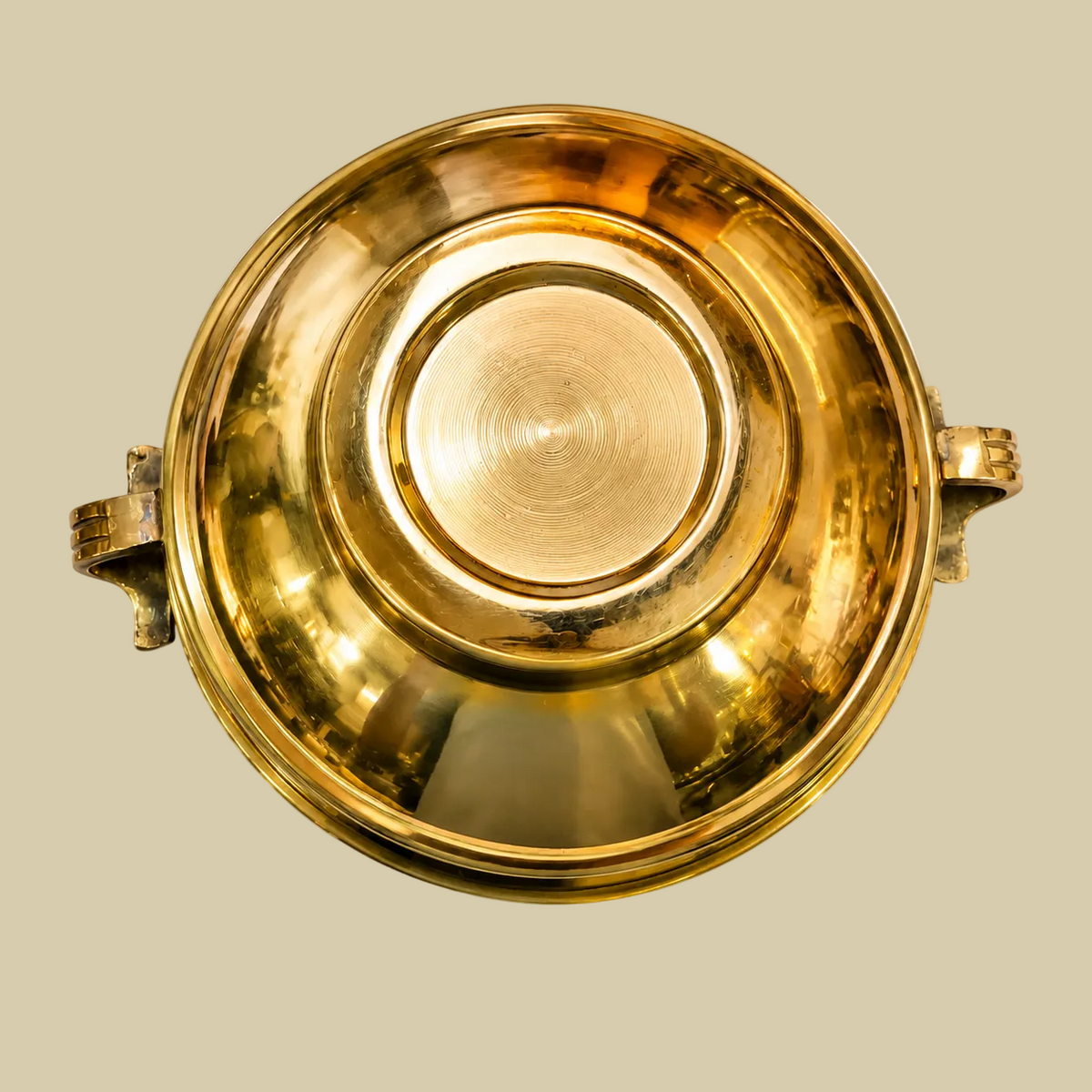 A polished golden plain brass Urli bowl with side handles, shown on a warm beige background. The bowl is reflective and has a warm golden hue under soft lighting.
