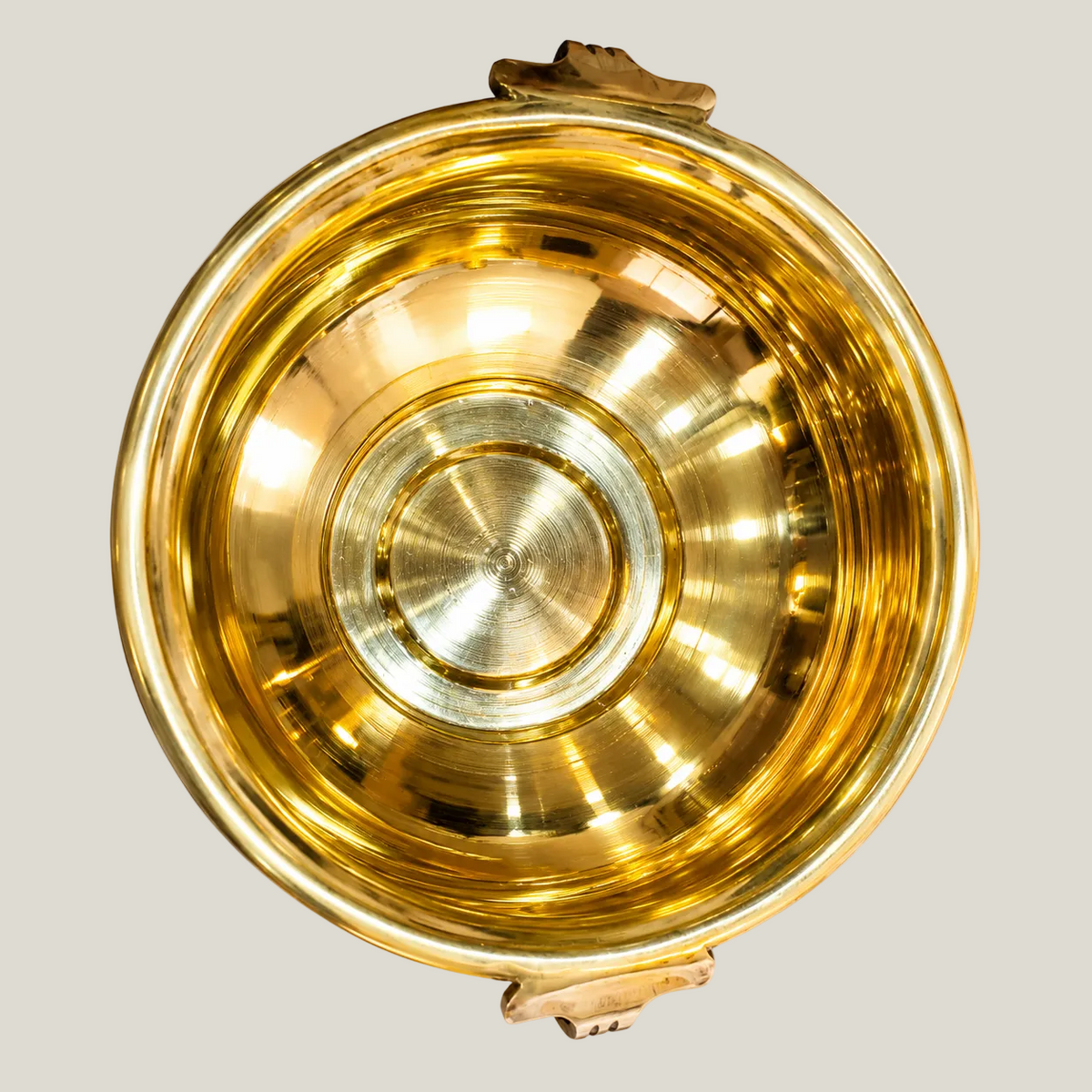 A polished golden plain brass Urli bowl with side handles, shown on a warm beige background. The bowl is reflective and has a warm golden hue under soft lighting.