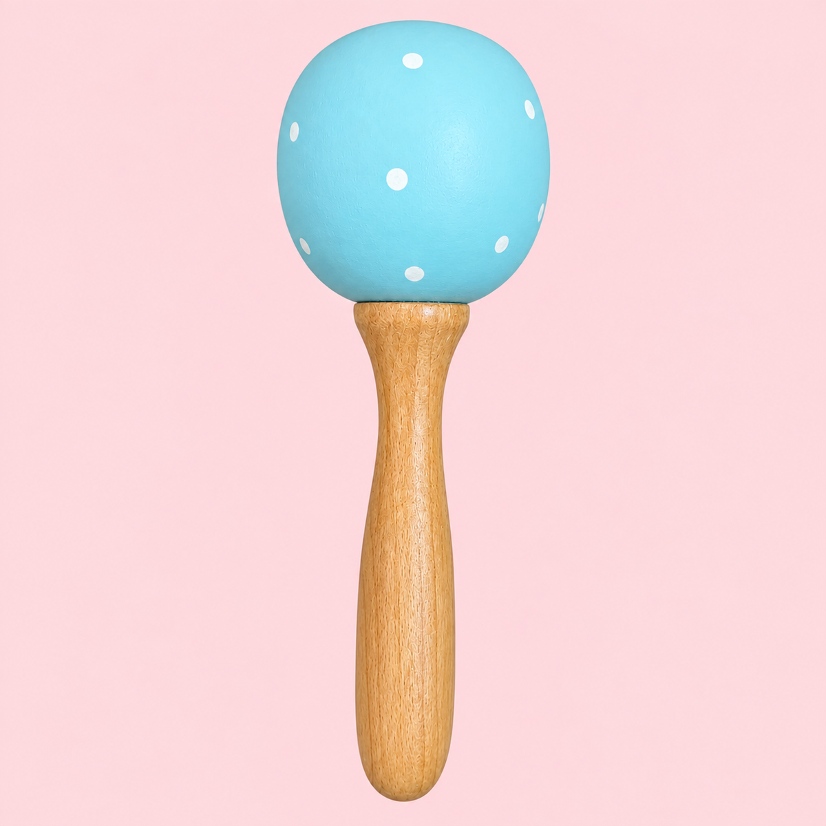 A baby toy maraca with a light blue wooden head decorated with white polka dots and a natural wood handle, set against a solid pink background.