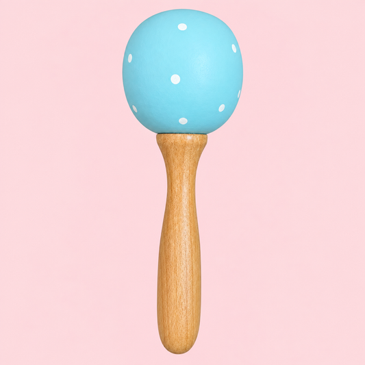 A baby toy maraca with a light blue wooden head decorated with white polka dots and a natural wood handle, set against a solid pink background.