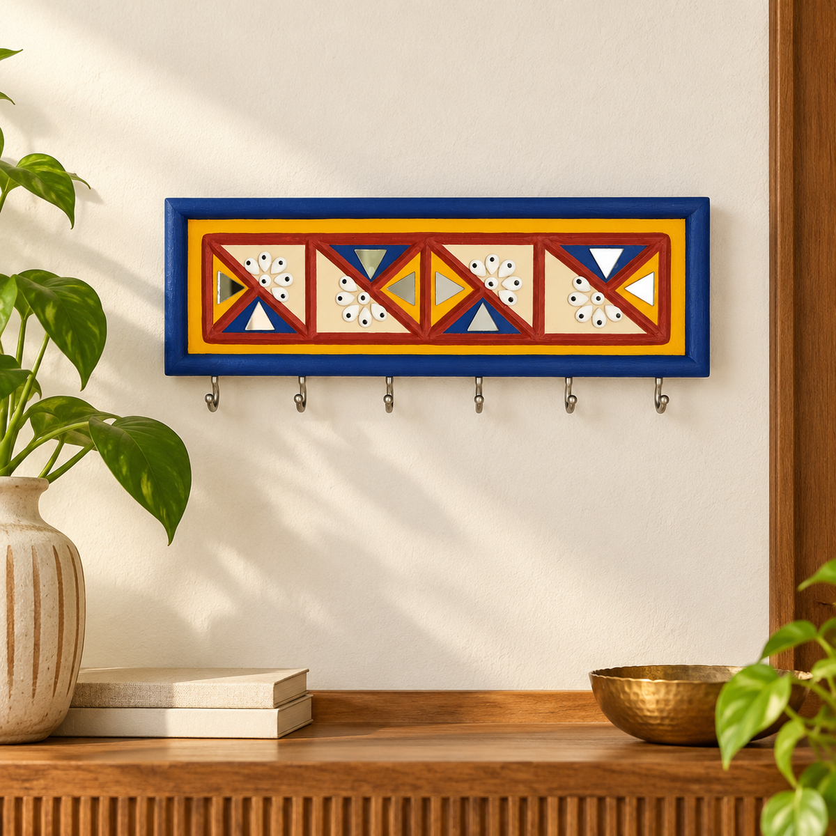 A rectangular decorative wall mounted key holder with five metal hooks, featuring a colorful Lippan art geometric pattern in red, blue, yellow, and cream, set against a plain off-white wall.