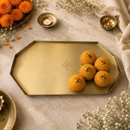 An octagonal brass gold-toned serving tray with a subtle floral engraving, styled with orange sweets on a beige fabric surface with decorative flowers and small brass oil lamps.