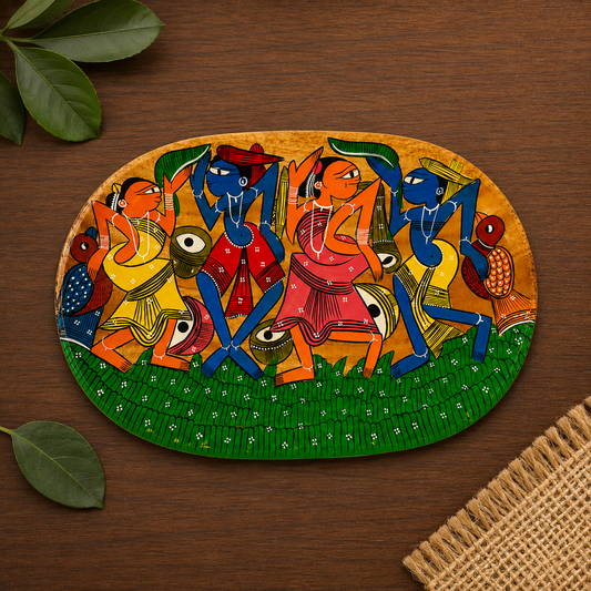 An oval hand-painted wooden serving tray featuring a vibrant Pattachitra art illustration of dancing figures, displayed on a dark wood tabletop with decorative green leaves and a woven mat corner.