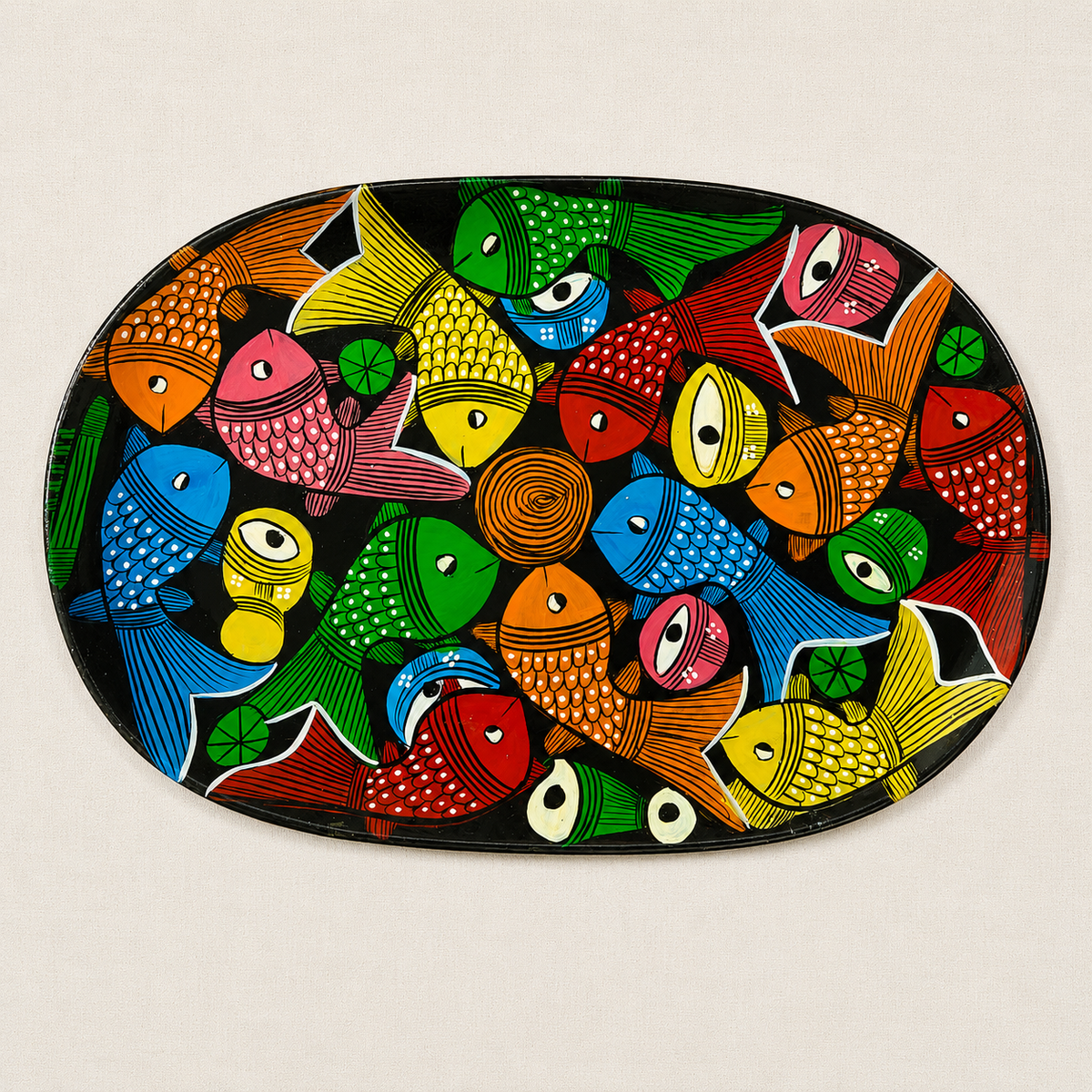 An oval decorative wooden serving tray featuring a vibrant, hand-painted Patachitra illustration of colorful fish on a black background, set against a light beige textured backdrop.