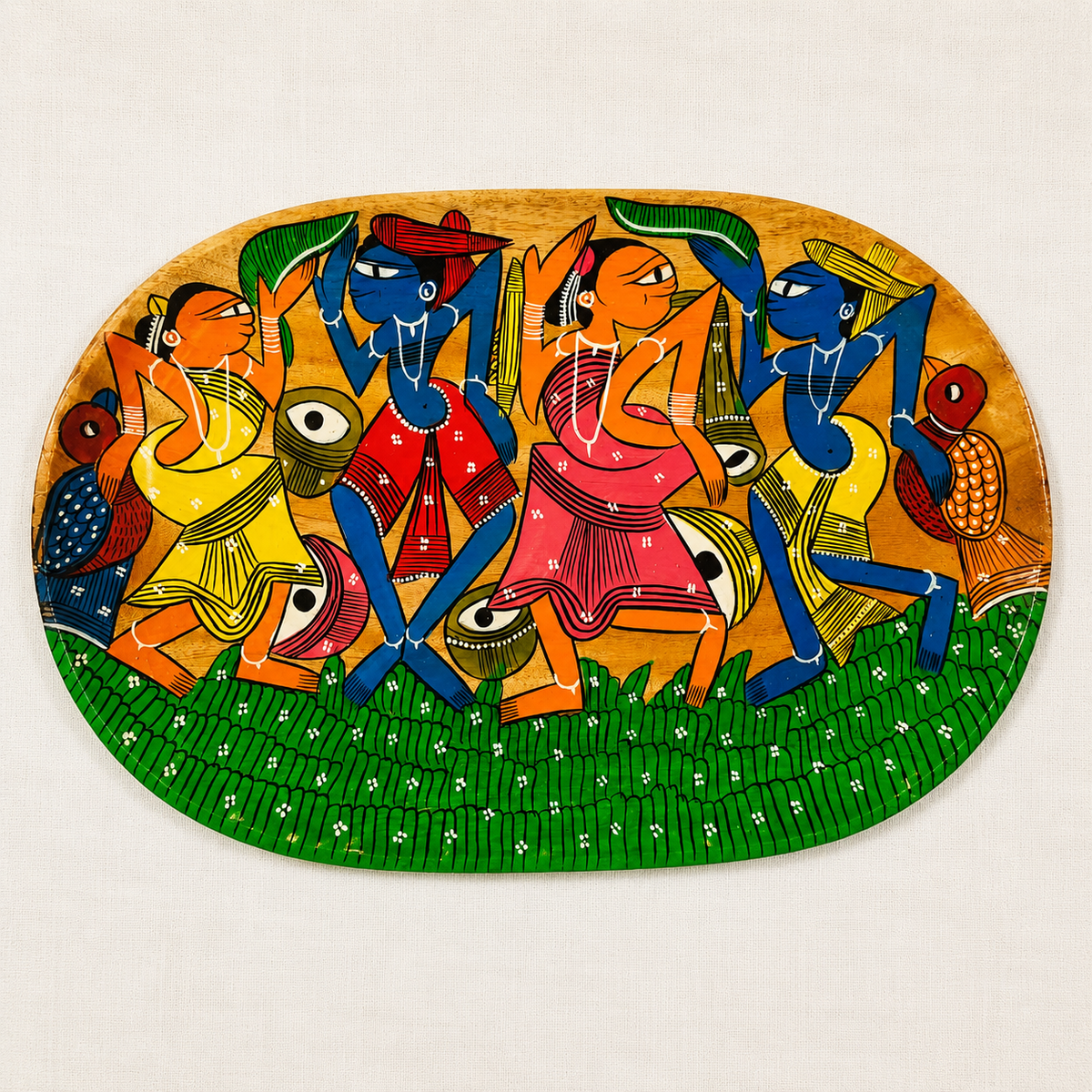 An oval wooden serving tray  with Patachitra painting depicting several stylized tribal dancing figures in bright yellow, red, blue, green, and orange on a tan background, shown against a light fabric backdrop.