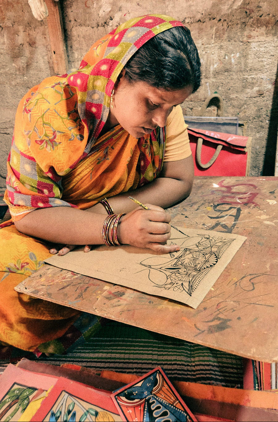 A woman in a colorful yellow and pink sari sits at a wooden table, engrossed Patachitra painting on paper. The setting appears rustic and workshop-like, with shelves and tools in the background.