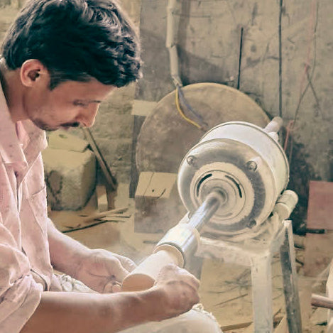 A man in a workshop sits on a metal chair, creating wooden handicraft. He wears a light pink patterned shirt and ripped blue jeans.