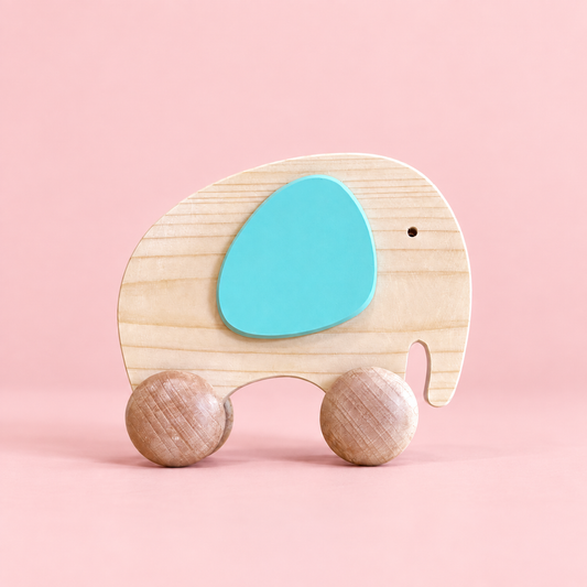 A wooden push n pull toy elephant with a natural light wood body, a bright turquoise blue painted ear, and two round natural wood wheels, set against a solid pink background.
